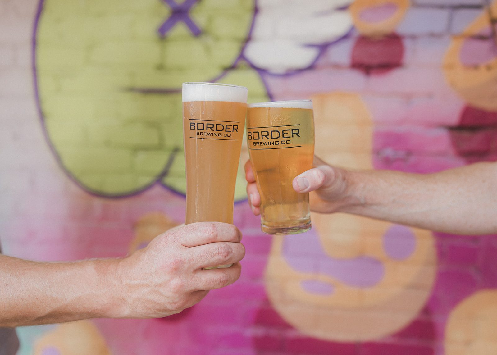 Two hands toasting branded Border Brewing pint glasses in front of a pink and lime graffiti mural