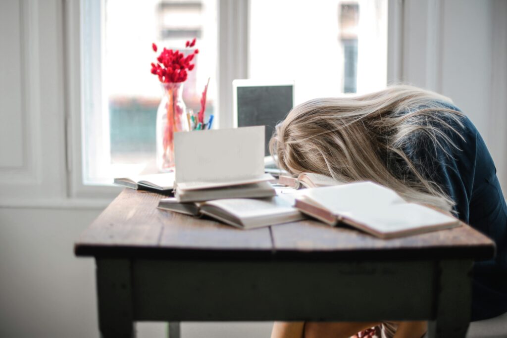 White woman falling asleep on her desk with books spread all over due to sleep deprivation.