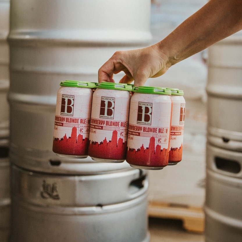 6 Pack of Strawberry Blonde Ale in front of some kegs in our Crossroads Art District location